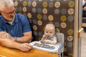 busy baby silicone placemat on travel highchair tray and baby chewing on busy baby 2-in-1 teether & training spoon with grandpa sitting next to them
