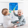 Two babies sitting on the floor playing with toys attached to the Busy Baby Original Mat which is stuck to the dishwasher.