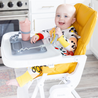 A baby sitting at a highchair with the busy baby original mat on the surface and the busy baby bottle bungee holding a cup in place with the baby holding the busy baby spoon from the busy baby utensil set