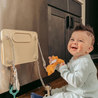 baby sitting in front of dishwasher with tan busy baby silicone placemat attached and toys attached to tethers baby smiling