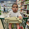 baby sitting in shopping cart with tan busy baby silicone placemat looped over covering shopping cart handle