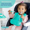 A baby sitting in a highchair at the kitchen counter wearing the Busy Baby Stop-Drop Bib+ Utensils holding the spoon with yogurt on it.