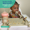 A baby sitting at a highchair with the Busy Baby Original Mat on the surface, eating from the Busy Baby Teething Spoon